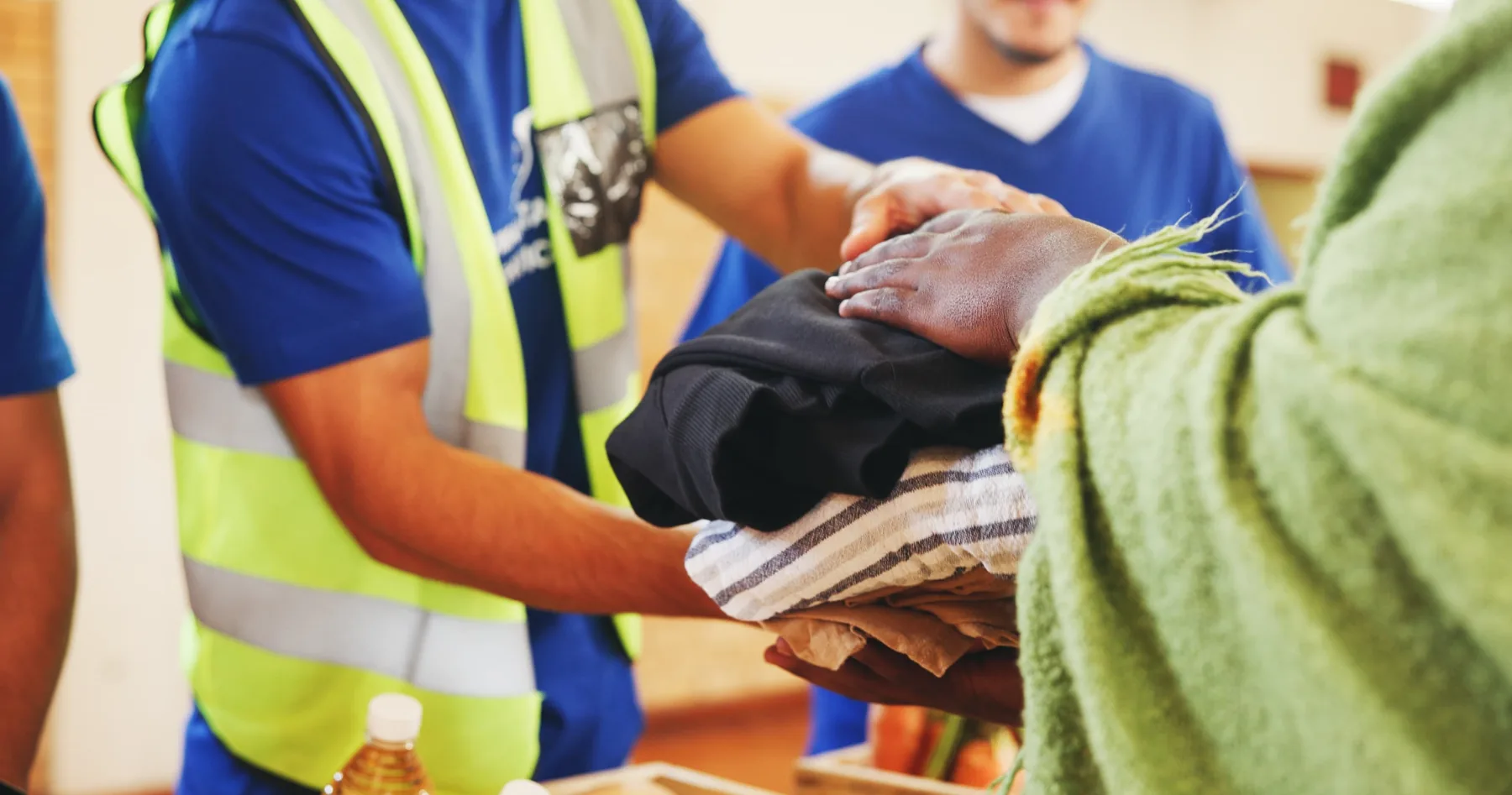 A relief worker in a high-visibility vest handing folded clothing and supplies to a person receiving aid