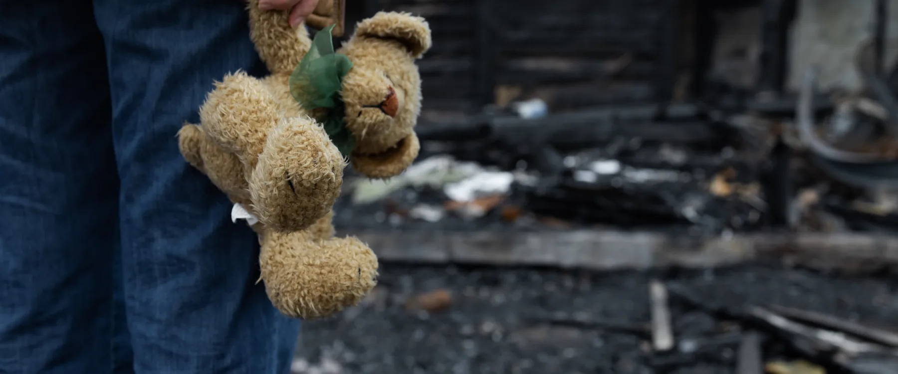 A person holding a child&rsquo;s teddy bear in front of a home destroyed by disaster