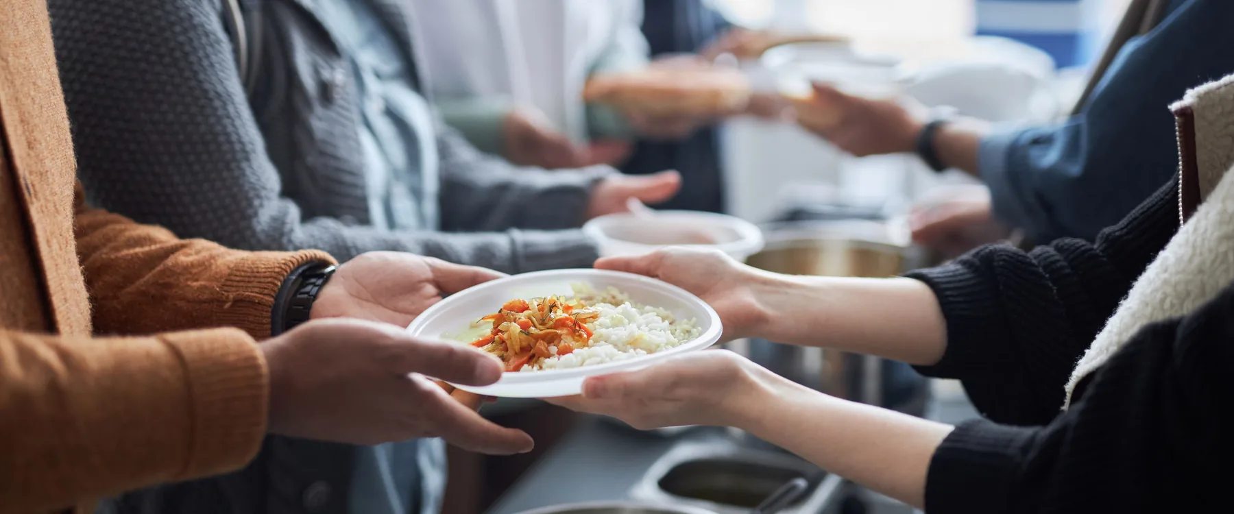 Hands passing a plate of hot food across a serving table to people waiting in line