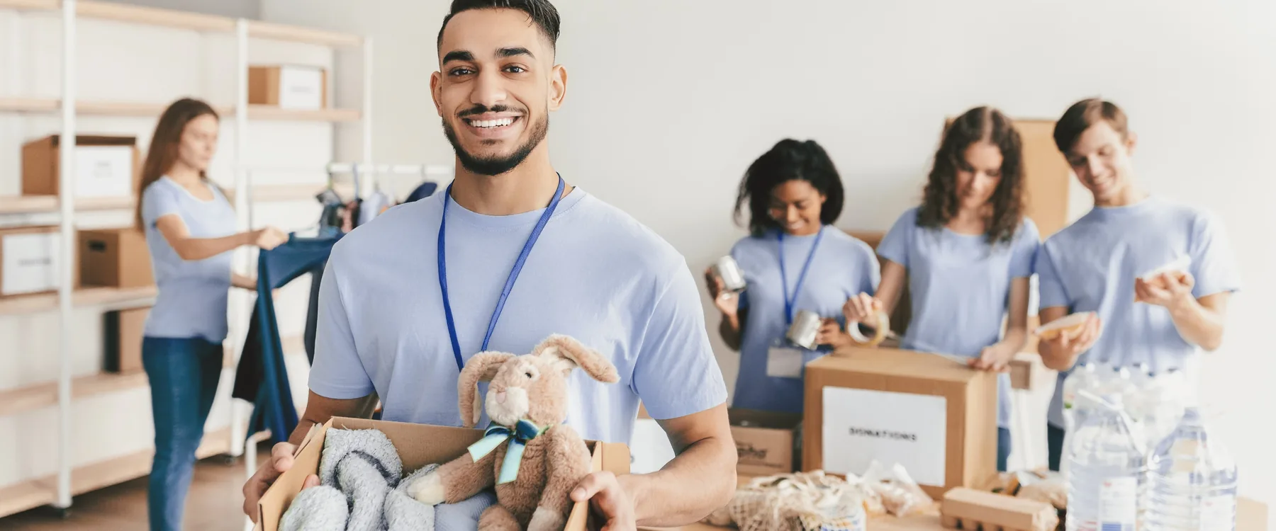 A smiling volunteer holding a box of donated blankets and toys while others sort supplies behind him